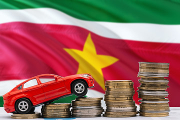 Suriname savings concept. Money for new automobile, toy car and coin piles standing on national flag background. Copy space for text.