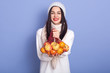© sementsova321 - Portrait of positive energetic model with black hair, smiling sincerely, having red lips, wearing hat, sweater and scarf, holding handbag with apples and mandarines in both hands. Vitamins concept.
