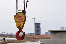 Large Yellow-red Crane Hook On Lifting Fears And Chains Hanging On It For Lifting Loads On A Background Construction Site, Resedental Building And Jib Crane