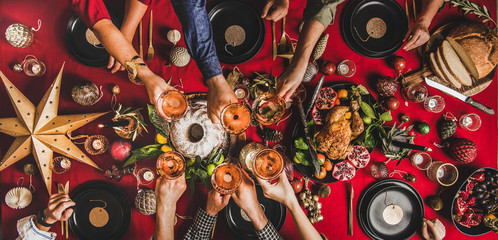 Wall Mural - Friends celebrating Christmas. Flat-lay of people clinking glasses with rose wine over festive table with red cloth with roasted chicken, bundt cake, fruit, decorations, top view. Winter holiday party