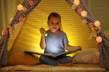 Canvas Print - Little girl with flashlight reading book in play tent at home