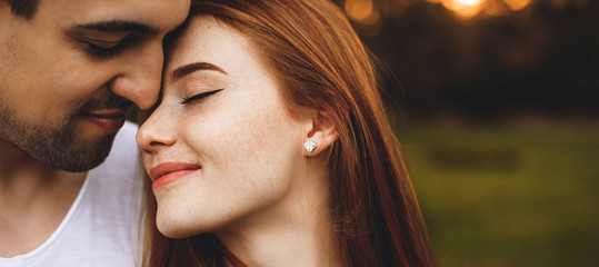 Wall Mural - Close up portrait of a beautiful young couple sitting face to face with eyes closed smiling before kissing while man is embracing her from back against sunset.