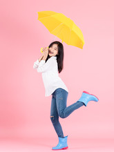 Asian Teenager Girl Holding Yellow Umbrella During Cold Rainy Day And Jumping On Pink Background.  Autumn Concept