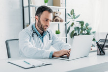 Positive Doctor Sitting At Workplace And Using Laptop
