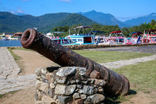 Close Up Of An Old Rusty Cannon In Front Of An Open Space With Grass, Colorful Boats And Rainforest Mountains, Historic Town Paraty, Brazil, Unesco World Heritage