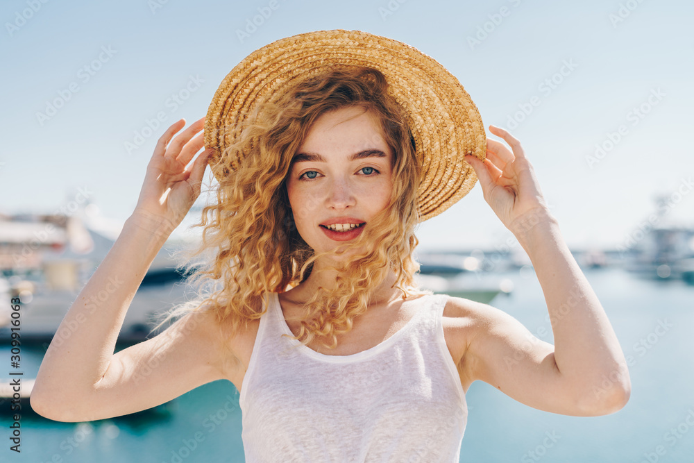 lovely girl with doll beauty holds a hat on her head with two hands on the background of ships and the ocean