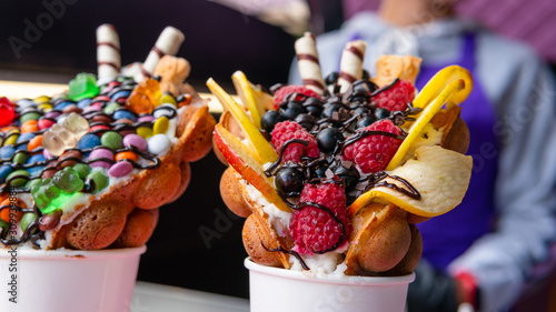 Fotografía Bubble waffles with raspberries, chocolate and marshmallow in white paper cups are sold on Open kitchen food festival event