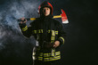 © alfa27 - portrait of professional and confident fireman holding hammer, wearing protective uniform and helmet on head, stand isolated in smoky background