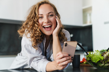 Wall Mural - Smiling young woman using smartphone in kitchen