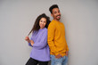 © timtimphoto - Pleasant looking smiling young dark skinned brunette couple leaning on backs of each other while standing over grey background, wearing colourful woolen sweaters