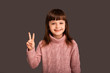 © Marharyta Hanhalo - Peace and love to everyone, cute pleasant brunette girl on a gray background shows a gesture two fingers up, kind and happy child