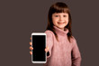 © Marharyta Hanhalo - Happy little girl on a gray background held out her hand forward while holding a phone and showing a mobile screen to the camera. Copy space. Digital technology