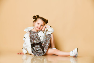 Smiling kid girl in leopard print dress, denim jacket is sitting on the floor leaning her head on her hand