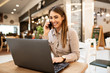 © Sanja - Cheerful attractive young caucasian brunette smiling and talking on her smart phone, She is also browsing on her laptop while sitting in a coffee shop.
