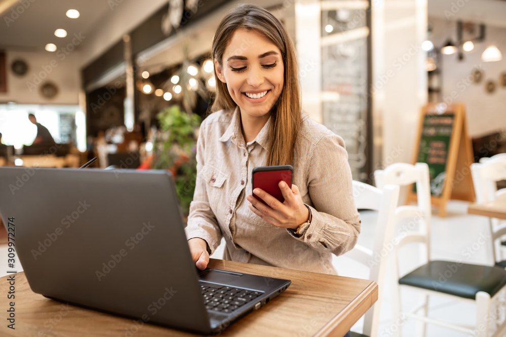 Attractive young caucasian businesswoman with brown hair smiling and looking at her smart phone while she is working on her laptop. She is sitting in a coffee shop. - obrazy, fototapety, plakaty 