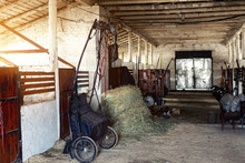 Old Empty Stables Barn At Horse Farm With Wooden Beams , Sulky Cart And Hay For Feeding Animals. Bright Sunset Sky Shining From Side Window. Countryside Village Rural Scene Background