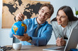 © LIGHTFIELD STUDIOS - attractive travel agent standing near smiling colleague pointing with pen at globe