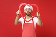 Shocked young bearded male chef cook or baker man in striped apron white t-shirt toque chefs hat isolated on red wall background. Cooking food concept. Mock up copy space. Holding big wooden heart.