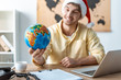 © LIGHTFIELD STUDIOS - selective focus of cheerful travel agent in santa hat holding globe while sitting at workplace