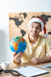 © LIGHTFIELD STUDIOS - selective focus of smiling travel agent in santa hat holding globe while sitting at workplace
