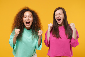 Two happy women friends european and african american girls in pink green clothes posing isolated on yellow orange wall background. People lifestyle concept. Mock up copy space. Doing winner gesture.