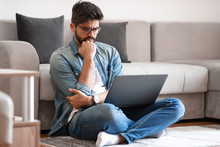 Confused Handsome Hipster Sitting With Legs Crossed On Floor In Living Room, Holding Laptop In Lap And Asking Himself About Bad Situation On His Bank Account.