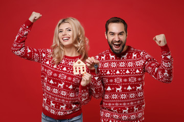 Wall Mural - Young couple guy girl in Christmas sweaters posing isolated on red background. Happy New Year 2020 celebration holiday party concept. Mock up copy space. Hold house bunch of keys doing winner gesture.