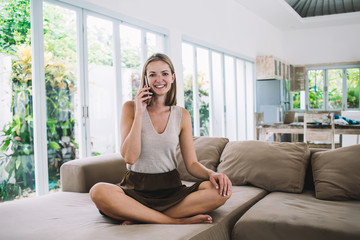 Cheerful woman talking on mobile phone and sitting on couch in apartment