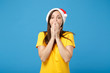 © ViDi Studio - Shocked young woman Santa girl in yellow t-shirt Christmas hat posing isolated on blue wall background. Happy New Year 2020 celebration holiday concept. Mock up copy space. Covering mouth with hands.