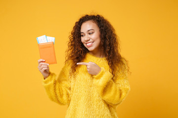 Wall Mural - Pretty young african american girl in fur sweater posing isolated on yellow orange background. People lifestyle concept. Mock up copy space. Pointing index finger on passport boarding pass tickets.