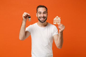 Smiling young man in casual white t-shirt posing isolated on orange background studio portrait. People sincere emotions lifestyle concept. Mock up copy space. Holding in hands house and bunch of keys.