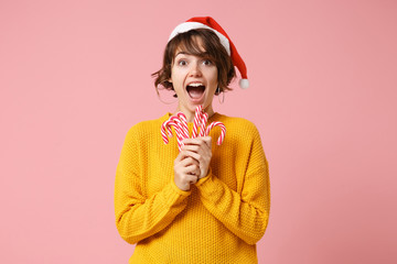 Shocked young brunette woman Santa girl in yellow sweater Christmas hat posing isolated on pink background. New Year 2020 celebration holiday concept. Mock up copy space. Holding striped lollipops.