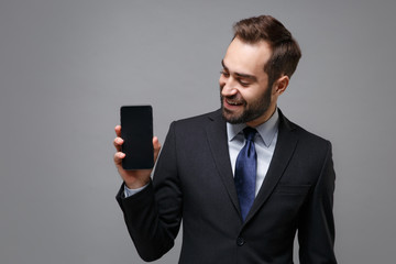 Wall Mural - Cheerful young business man in classic suit shirt tie posing isolated on grey background. Achievement career wealth business concept. Mock up copy space. Holding mobile phone with blank empty screen.