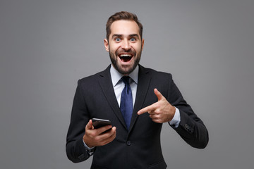 Excited young business man in classic black suit shirt tie posing isolated on grey background. Achievement career wealth business concept. Mock up copy space. Pointing index finger on mobile phone.