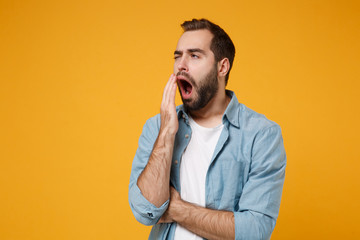 Tired young bearded man in casual blue shirt posing isolated on yellow orange wall background studio portrait. People emotions lifestyle concept. Mock up copy space. Yawning, covering mouth with hand.