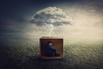 Surreal concept with a scared guy shelters inside a cardboard box. Introvert man caged by own fears as a thunderstorm cloud trapped him under the rain. Mysterious storm as emotional crisis symbol.