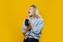 Playful Grimacing Blonde Woman Hairdresser In A Jeans Belly-tied Shirt Is Fooling Around, Singing In A Hair Dryer, Using It Like Microphone Over Yellow Background.
