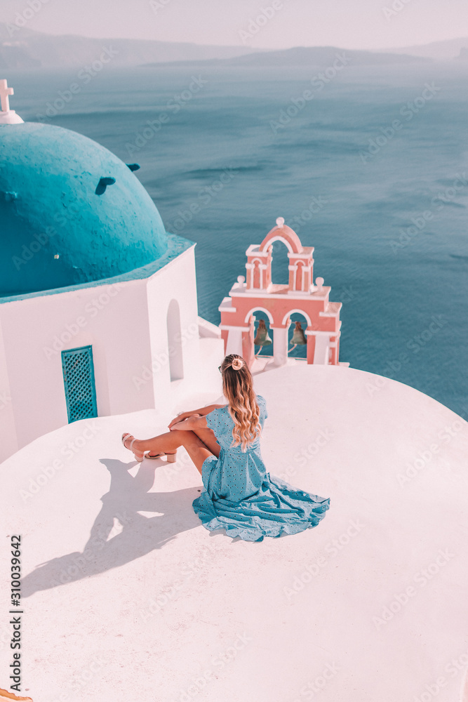 Young woman with blonde hair and blue dress in oia, santorini, greece with ocean view and churches - obrazy, fototapety, plakaty 