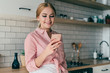 © GalakticDreamer - Adult attractive woman in casual clothes surfing in internet using phone standing in kitchen with white tile on wall smiling