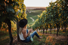 Young Blonde Woman Relaxing In The Vineyards In Summer Season. Girl Sitting Near The Bottle And Holding Glass White Wine. Outdoor Farmer Countryside Style
