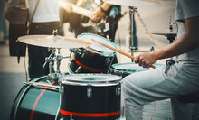 A Drummer In Gray Sportswear Plays Wooden Sticks On A Drum Kit, Setting The Rhythm To A Street Band Of Musicians Illuminated By The Sun.