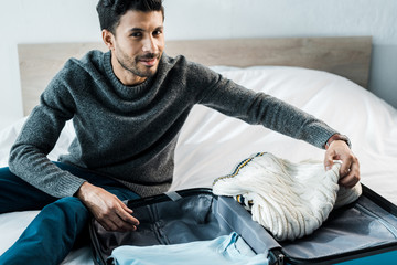 handsome and smiling bi-racial man in sweater packing travel bag in apartment