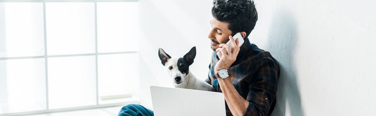 panoramic shot of handsome bi-racial man talking on smartphone and holding jack russell terrier