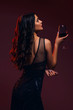 Glamour caucasian brunette woman in evening black dress on a neutral background. She pose sensually with glass of wine. Retro fashion mood.
