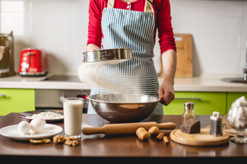 Cook housewife making cookies at home on colorful kitchen. Woman sifts flour through sieve at the table with ingredients