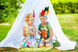 3 years old girl and 5 years old boy with feathers on head sit near white tent with tambourine and maracas in hands. Children pretend to be indian. Indians game in summertime. Children play music