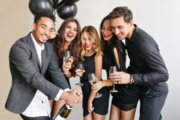 Wall Mural - Handsome african man with beard opens bottle of champagne to celebrate something with friends. Indoor portrait of blissful young people spending time together at birthday party.