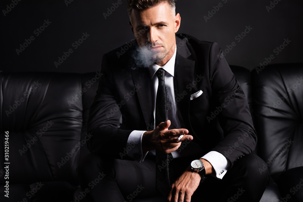 Handsome businessman smoking cigarette on sofa isolated on black Stock  Photo