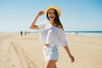 girl dynamically walking along the beach past the camera, laughs, holding her hat with her hand