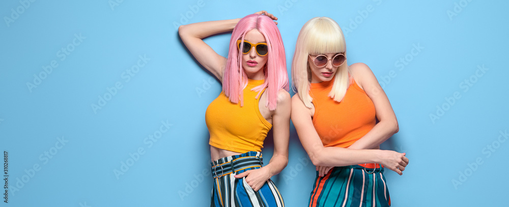 Two Young carefree Woman in stylish sunglasses, striped pants. Beautiful fashionable model girl in trendy summer outfit. Graceful friends with fashion dyed hairstyle, make up on blue. Creative banner - obrazy, fototapety, plakaty 